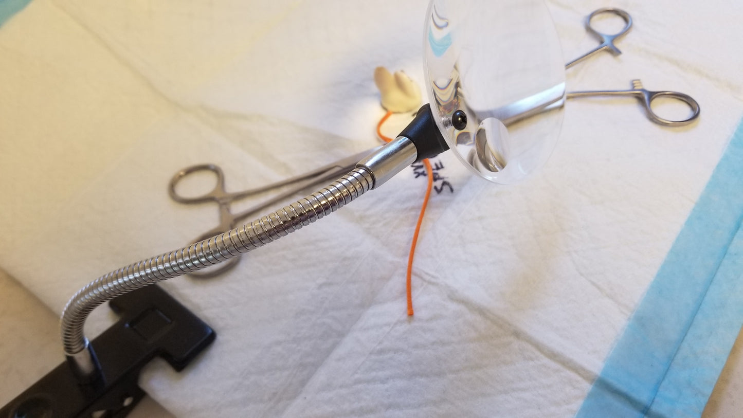 Close up view of Clip on Magnifier, Adjustable. Has a 2cm diameter inset lens on a metal flexible neck looking over a laboratory work station that includes lab tools and lab mouse. Clip on Magnifier available at National Supply Company LLC.