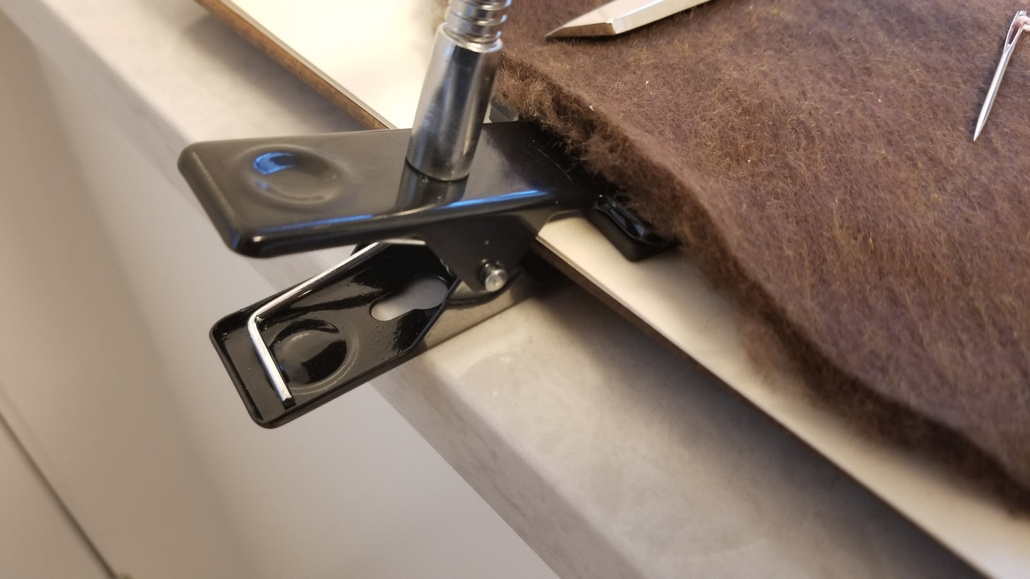 Hands Free Clip on Magnifier. An up close view of a metal black clip or clamp at the end of a metal stem neck magnifying glass securely attached to clipboard with craft supplies such as felt material, scissors, and sewing needle present. Get Hands Free Magnifier through National Supply Company LLC.