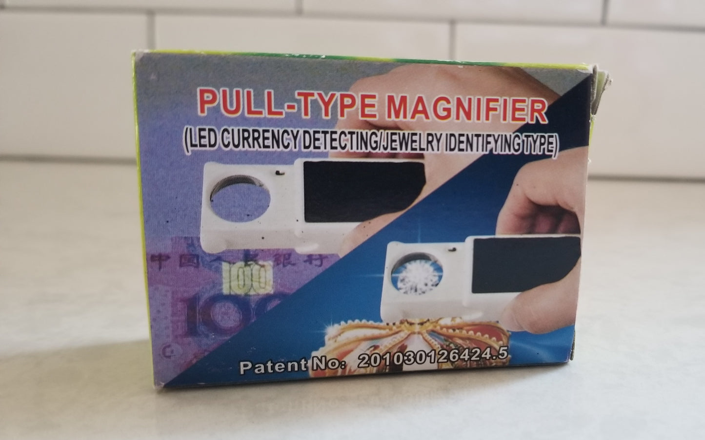 Pull Type Magnifier LED Currency Detecting/Jewelry Identifying Type. Box label shows Magnifier with UV type light over cash money for detection and bright white LED light over a gemstone for jewelry identification. Counterfeit Detector Tool and a Jewelry Identifier Tool. Product available at NSC LLC.